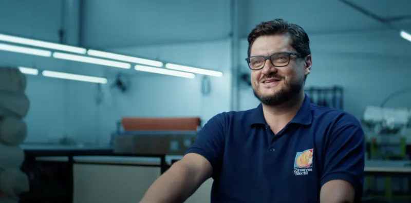 Empresário de camisa polo azul sorrindo no chão de fábrica de uma indústria têxtil